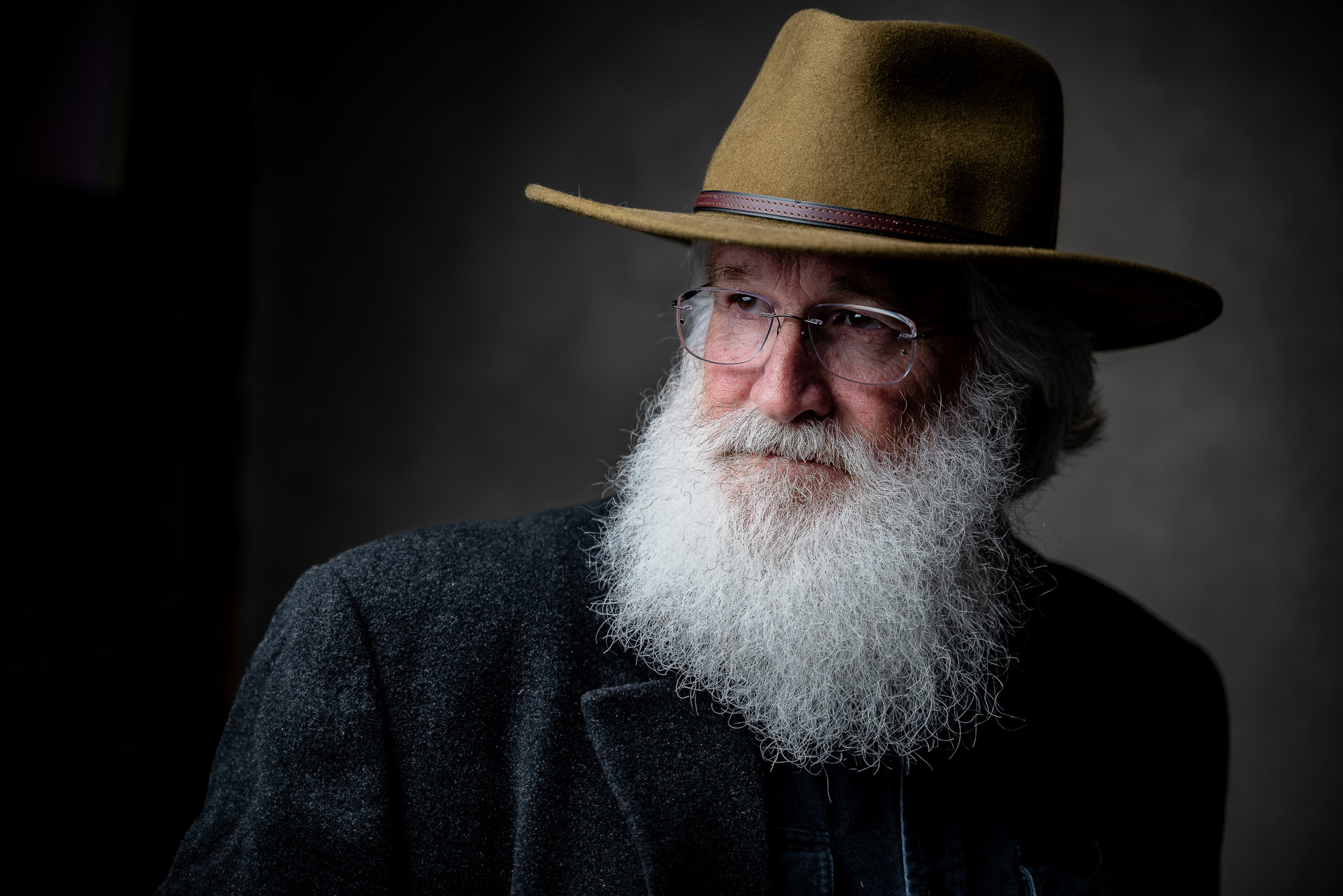 A portrait of a man with a long white beard and glasses, wearing a black jacket and brown hat. The image has a dark and mo...